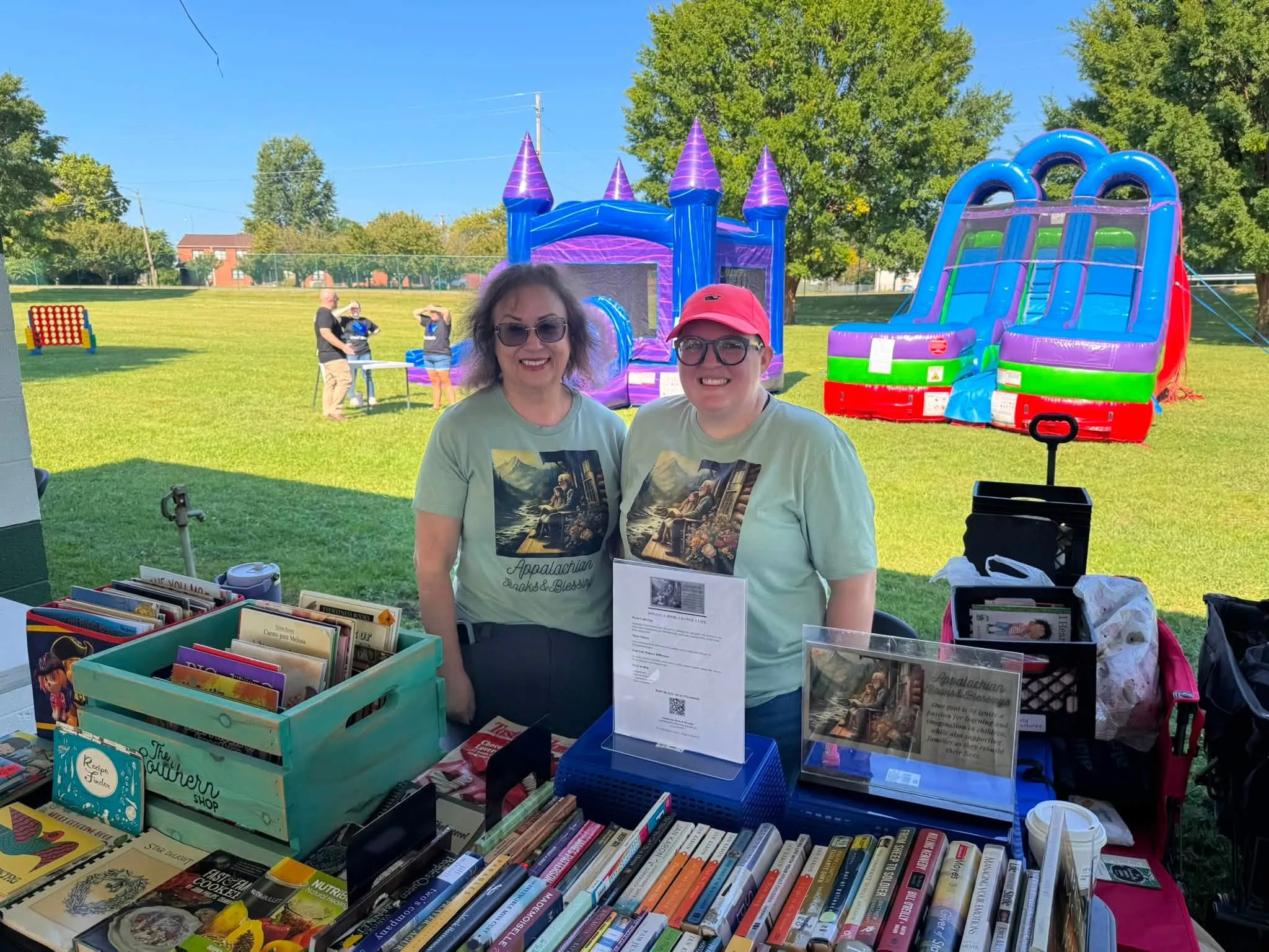 Two smiling volunteers in sage green Appalachian Books & Blessings shirts stand behind tables and crates filled with books at an outdoor community event.